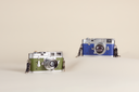 Two vintage-style compact cameras on a warm beige studio background. The camera on the left is olive green with silver metal details, and the camera on the right is blue with silver metal details.